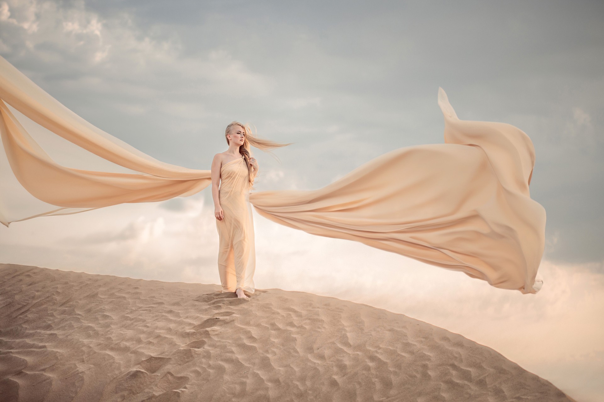 A blonde girl in a beautiful beige dress stands on a dune in the desert. the dress flutters beautifully in the wind. sunset.