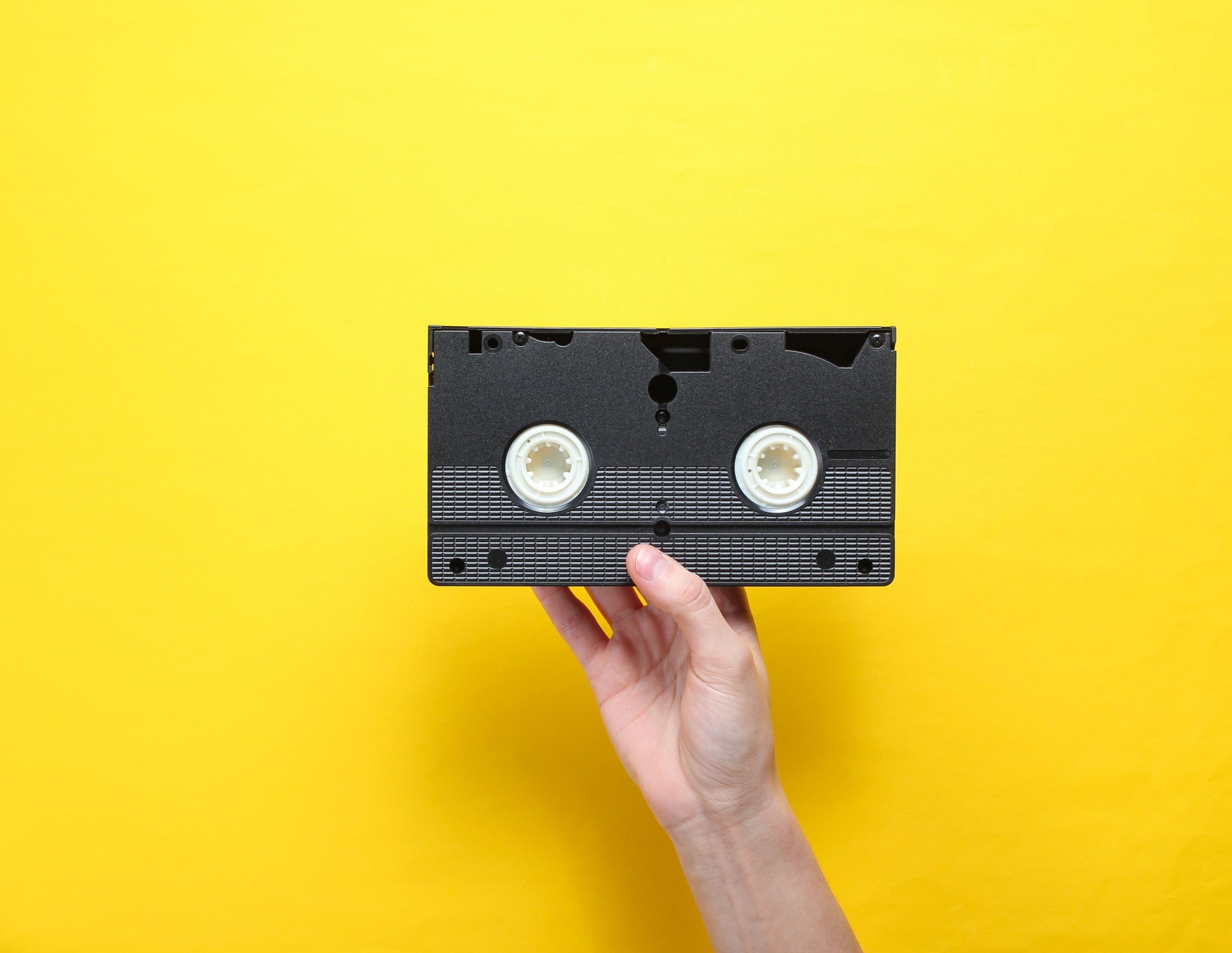 Female hand holds video cassette on yellow background. Retro style, pop culture, minimalism, top view